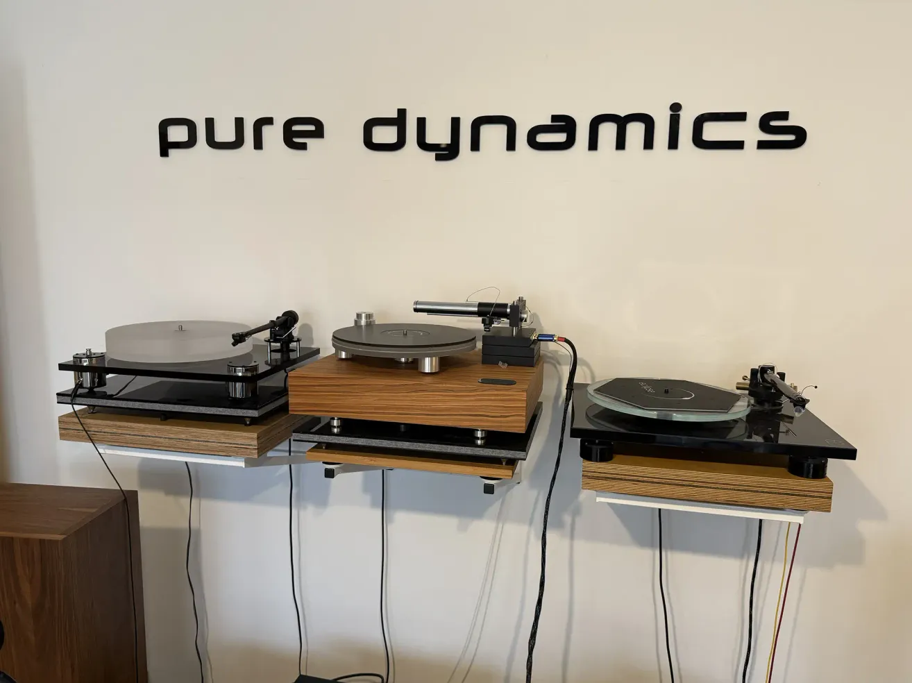 Three turntables are mounted on a white wall with the word Pure Dynamics. The turntables have wooden bases and are connected with wires. The turntables are connected to the wall with a bracket.