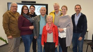 Five adults, including three women and two men, are smiling and standing together in a room. They are wearing casual clothes and seem to be posing for a photo. Behind them, there is a monitor with a logo.