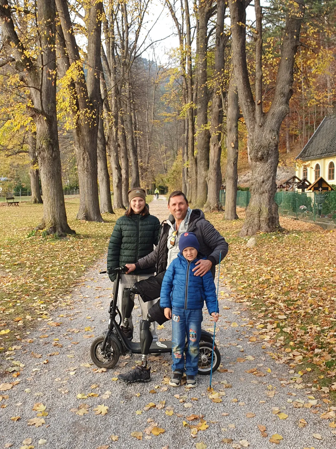 Eine dreiköpfige Familie steht auf einem Weg in einem Park. Ein Mann ist auf einem Roller mit einer Prothese. Eine Frau steht neben ihm. Ein junger Junge steht zwischen ihnen. Sie alle lächeln.