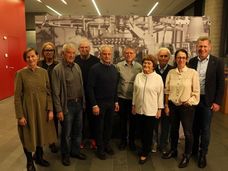 A group of people are standing in front of a large photograph of industrial machinery. They are all smiling and appear to be posing for a photo.