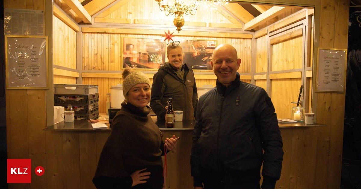 Three people stand in a cozy cabin, smiling, with a wooden wall, a counter, and a chandelier. A bottle is on the counter.