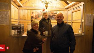 Three people stand in a cozy cabin, smiling, with a wooden wall, a counter, and a chandelier. A bottle is on the counter.