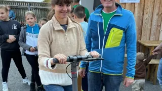 A man and woman stand with a scooter in front of a building. Behind them are children and adults, with one child reaching for something on a table. The building has windows and a sign.