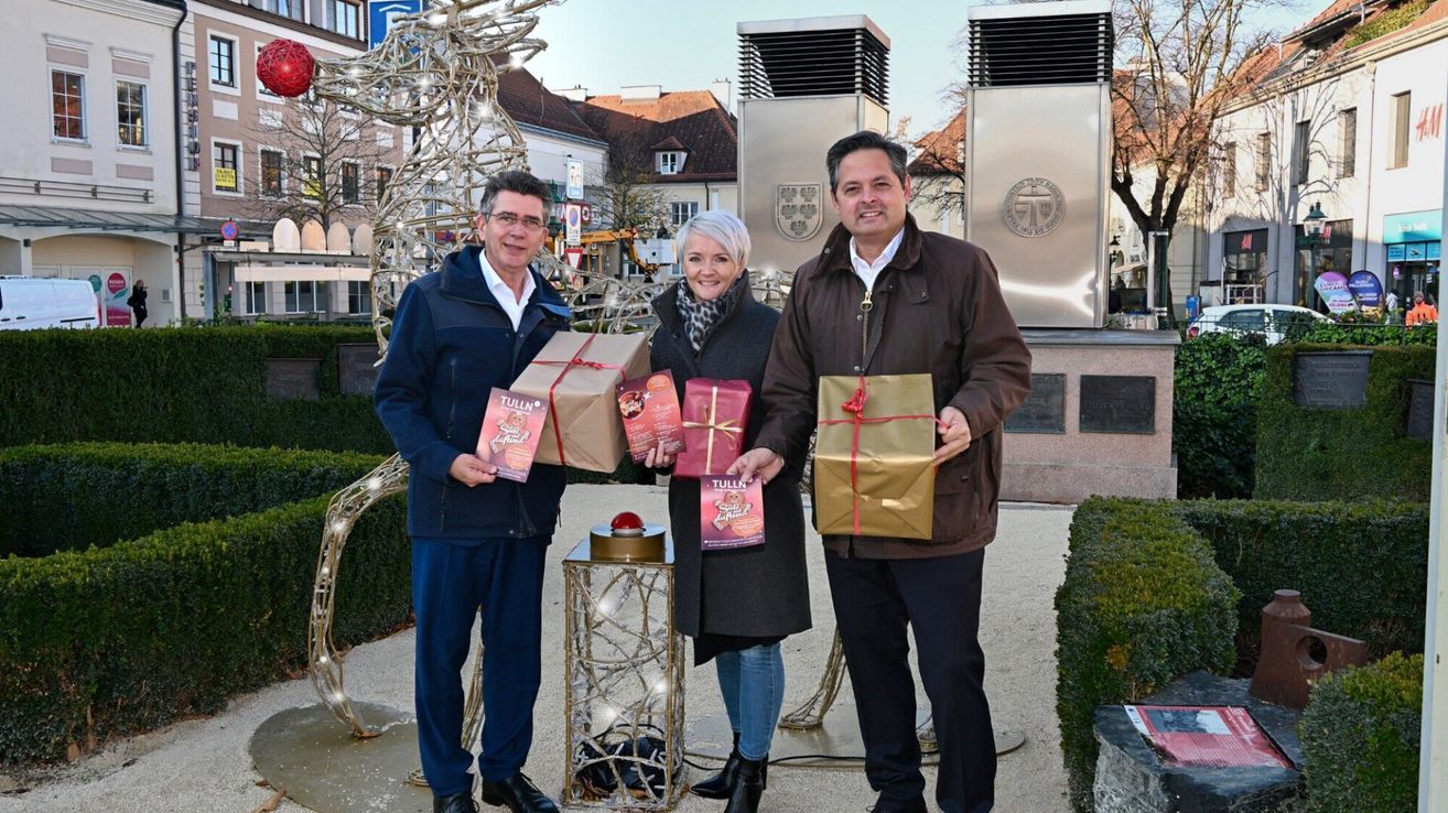 Three people stand in front of a Christmas tree, holding gifts. The man on the left wears glasses and a blue jacket. The woman in the middle wears a scarf and holds a gift. The man on the right wears a brown jacket. Behind them is a silver monument.