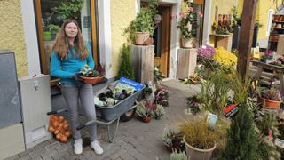 A woman stands in front of a shop, holding a wheelbarrow filled with potted plants. The shop has wooden boxes, plants, and flowers. The woman wears a blue top and white sneakers.