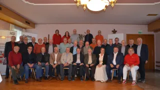 A group of people wearing formal attire sitting in chairs in a room with a chandelier hanging from the ceiling.