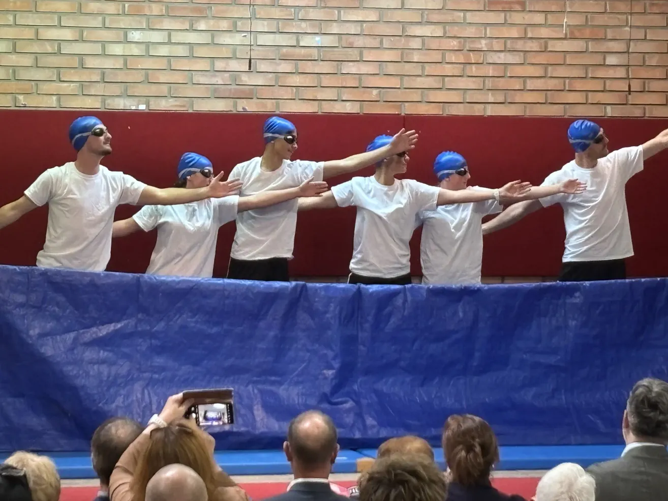 Five individuals in swimming caps stand behind a blue barrier, facing a crowd. The audience, seated in front, watches attentively. One person in the crowd holds a camera.