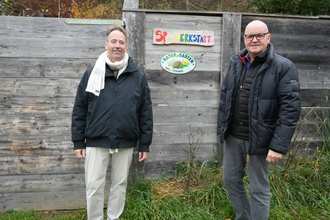 Two men stand next to a wooden fence with a sign that reads 'SP LEHRSTATT' and a circular emblem below it. They are dressed in black jackets and pants.