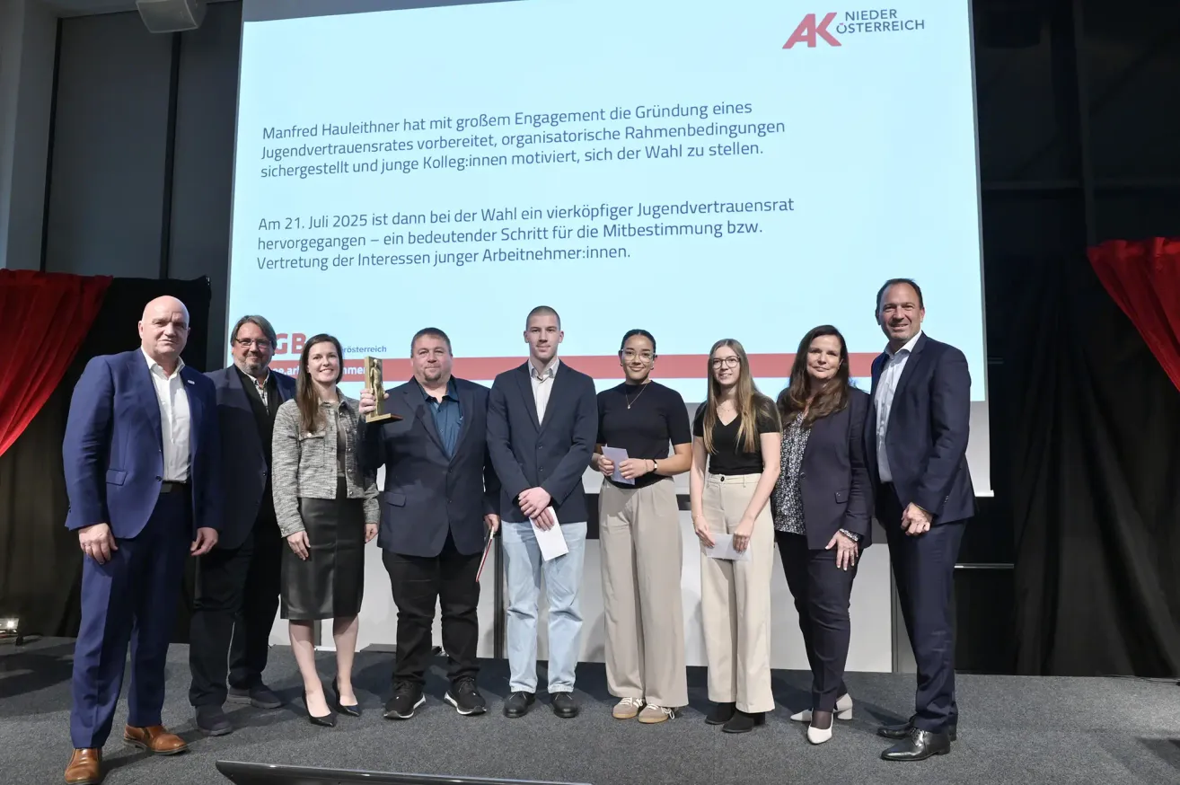 A group of people stands on a stage holding papers and a trophy, with a screen behind them displaying a message about the establishment of a youth trust committee.
