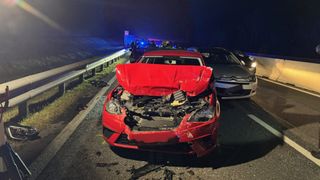 A red car is wrecked on a highway at night. The hood is open, showing the engine. Behind it, a gray car and emergency vehicles are present.