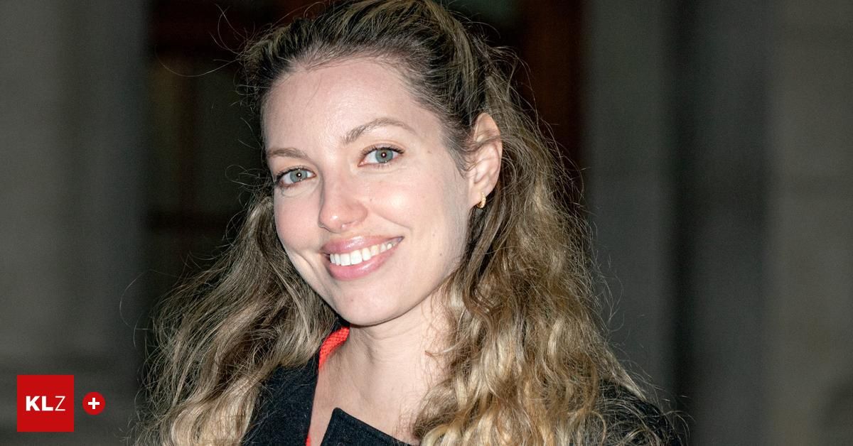A woman with curly blonde hair, wearing a black top, smiles at the camera. Her eyes are blue, and she has a mole on her right cheek.