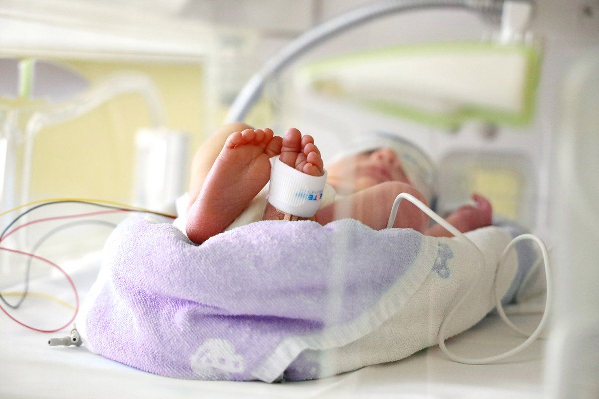 A newborn baby lies in a hospital bed, feet bandaged with a medical tag. The baby is wrapped in a purple and white blanket.