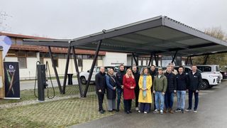 A group of people pose under a carport with electric car chargers, two white SUVs parked behind them, and a building labeled 'Informationszentrum' in the background.