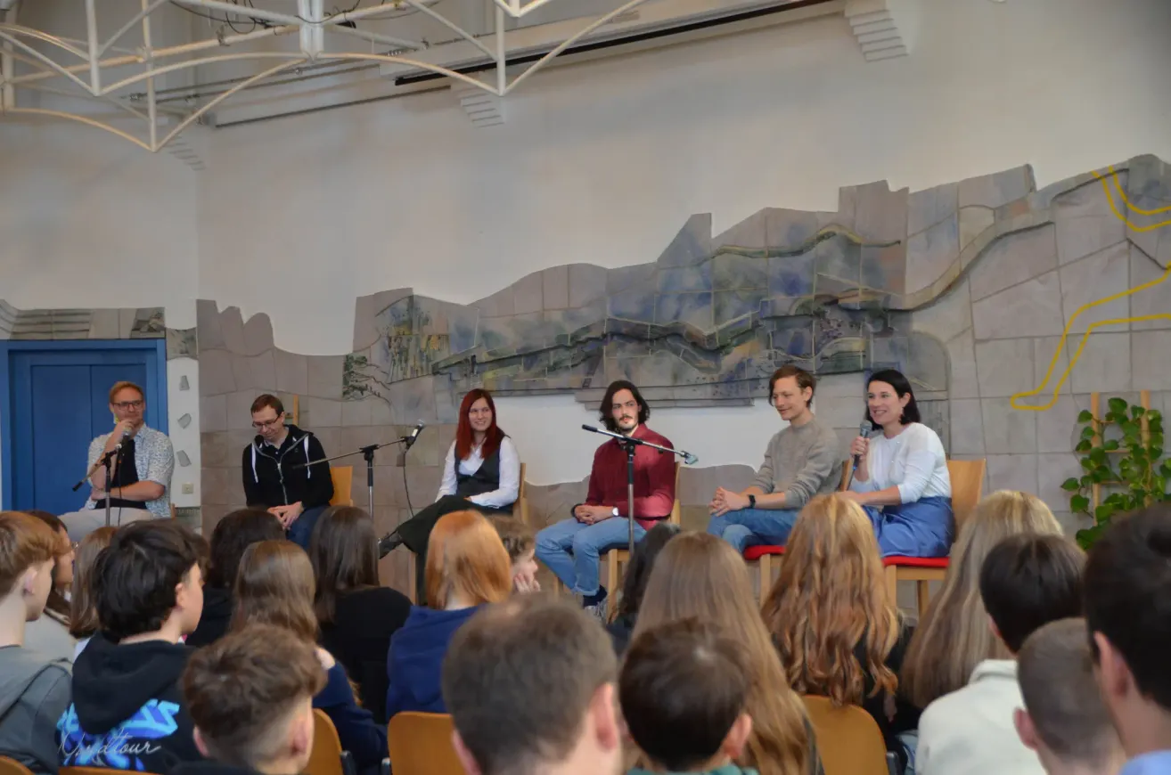 A group of people is seated in chairs in front of a large mural. Four individuals are positioned on a stage with microphones, likely engaged in a discussion or presentation. The audience is seated in front of them, attentively listening.
