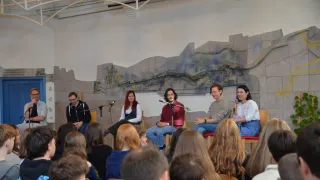 A group of people is seated in chairs in front of a large mural. Four individuals are positioned on a stage with microphones, likely engaged in a discussion or presentation. The audience is seated in front of them, attentively listening.