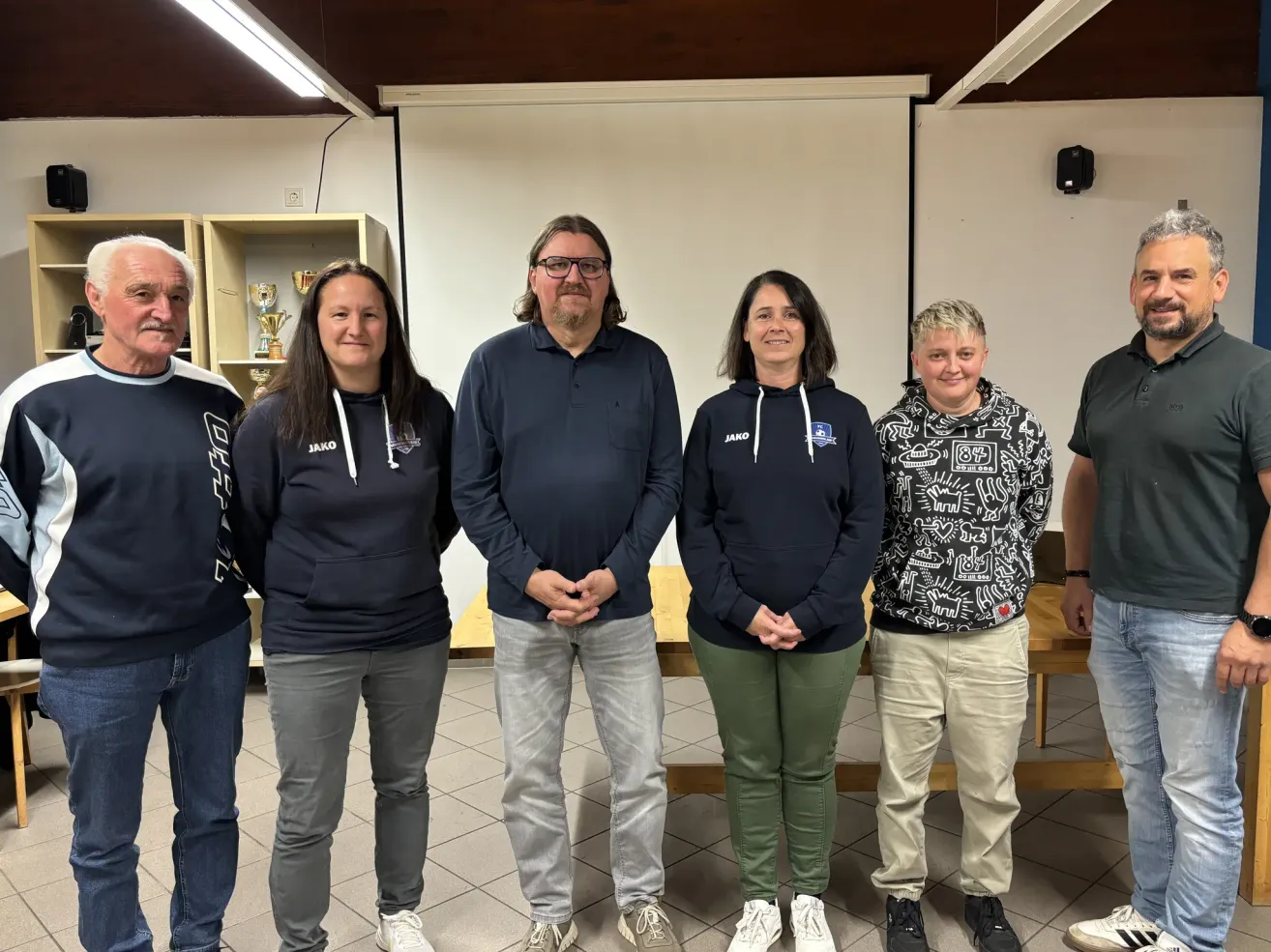 Five individuals stand together in a room with a tiled floor. Three of them wear navy hoodies, one in a patterned hoodie, and one in a polo shirt. Behind them, a table and a whiteboard are visible.