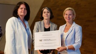 Three women stand smiling and holding a certificate. The woman on the left wears a striped shirt, the middle one a blazer, and the right one a white blazer. The certificate is white with black text and a logo.