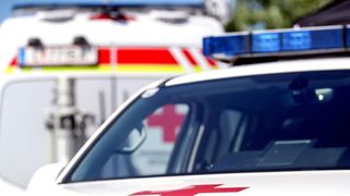 A white emergency vehicle with red crosses and blue lights on top is parked in front of a blurry background.