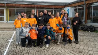 A group of people in orange shirts are posing for a picture in front of a building. There are also people in black clothing. Some are wearing glasses, and some are wearing hats.