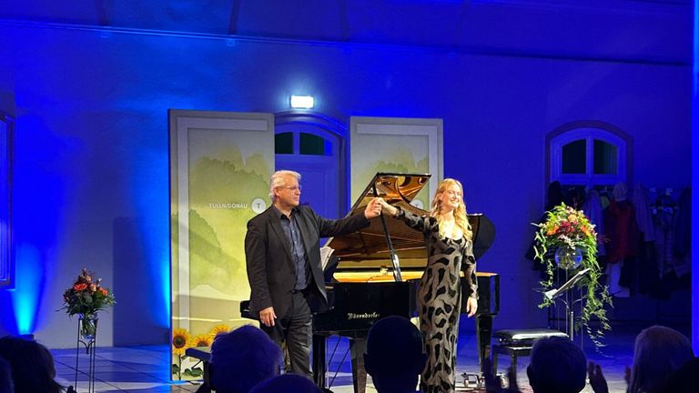 A man and a woman are standing in front of a grand piano on a stage. The man is holding the woman's hand. Behind them is a blue wall with a picture of sunflowers. The audience is watching them.