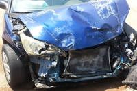 The front of a blue car shows significant damage with a crumpled bumper, a broken headlight, and a damaged radiator grill. The car is likely involved in a serious accident.