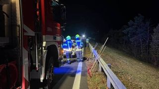 Two emergency workers are on the side of the road at night, one wearing a helmet and the other wearing gloves. A fire truck is parked nearby.