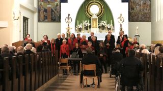 A choir dressed in red stands in a church, singing in front of a seated audience, near a decorated altar and religious artwork.