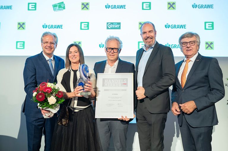 Five people stand together, smiling, with a large certificate in the center. The woman on the left holds a bouquet of flowers. The man on the right holds the certificate. Behind them, logos are displayed on a white backdrop.