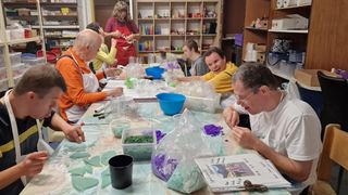 A group of people are gathered around a table, engaged in a crafting activity. Various materials and tools are spread across the table, including a blue bowl and clear plastic containers. One person is wearing glasses and a white shirt.