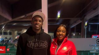 A man and a woman are standing close together, smiling for a photo. The man wears a white cap and a black hoodie with a logo. The woman is in a red jacket with text and a logo on the chest. They are in a room with a glass wall and a lamp above.