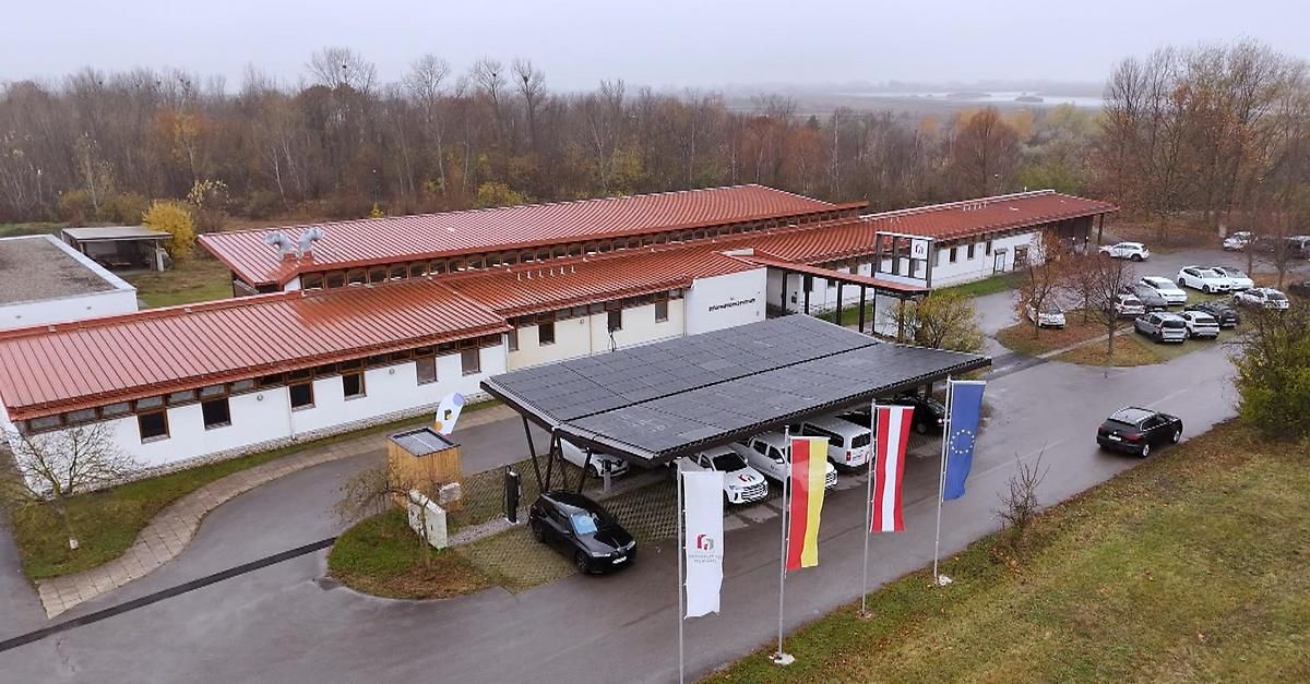 An aerial view of a modern building with a red roof and solar panels. Cars are parked under a canopy with flags and a small wooden structure.