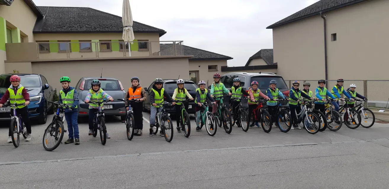 A group of children and young people in reflective vests and helmets stand with their bikes in a parking lot near buildings and cars.