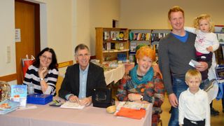 A group of people sit at a table inside a room, possibly a library. The man and woman wear smiling faces. They have books, a calculator, and other items on the table. Behind them are shelves filled with books and a sign on the wall.