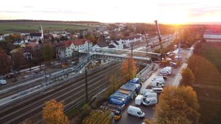 An aerial view of a construction site with workers, a bridge, a train track, and vehicles. A crane is positioned over the bridge.
