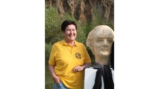 A woman in a yellow shirt poses with a stone bust of a man. The bust is wearing a scarf and has a serious expression. The woman is smiling.