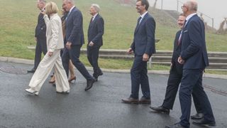 A group of formally dressed individuals, including men and women, walk together on a paved road with a grassy hill in the background.