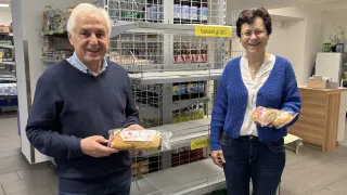 An older man and woman are standing in a bakery, smiling. The man holds a loaf of bread, and the woman holds a pastry. Behind them is a shelf with bottles and a yellow sign.