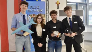 Five young people pose for a photo in front of a banner advertising Flug Technik. The two men on the ends hold a model airplane and a megaphone. The two in the middle hold what looks like a piece of technology.