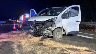 A white van with a damaged front and open doors is on the roadside at night. Emergency vehicles and cones are nearby.