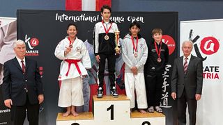 Five young athletes stand on a podium, wearing medals. They are dressed in martial arts uniforms, and the backdrop features a banner reading 'International Vienna Open'. The boy in the middle holds a trophy.