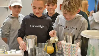 Four boys are preparing drinks at a bar counter. One boy in a Calvin Klein hoodie is pouring a drink, while another boy in a Tommy Hilfiger hoodie watches. They are surrounded by various cups, a glass pitcher, and straws.