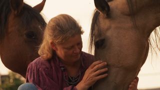 A woman with short blonde hair is gently touching a horse's face. The horse has its eyes closed, and another horse is standing behind the woman.