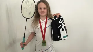 A young girl is holding a badminton racket and a backpack. She has a medal around her neck and is smiling.