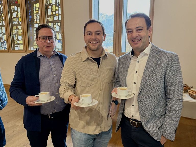 Drei Männer, zwei mit Kaffeetassen, stehen lächelnd vor einem Buntglasfenster.