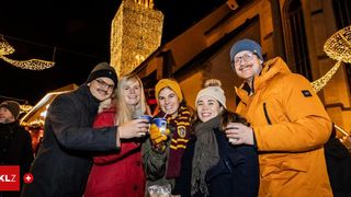 Five individuals, all smiling, pose for a photo at night, each holding cups and wearing winter clothing, in front of a lit-up tower.