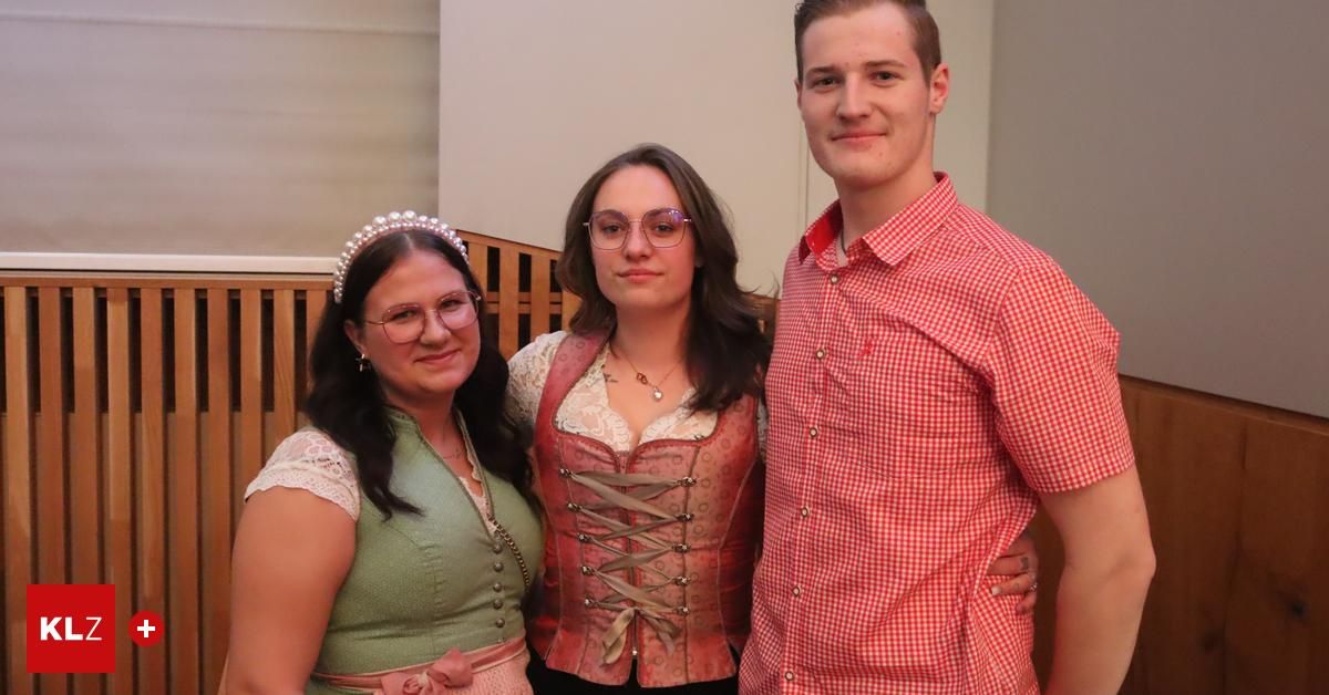 Three individuals, two women and one man, stand together. The women wear traditional dresses and the man wears a red and white checkered shirt. They are smiling.