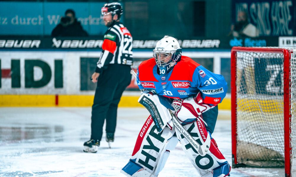 An ice hockey player wearing a blue and red jersey with the number 30 kneels in front of the goal. Behind him is a referee, and a person takes photos.