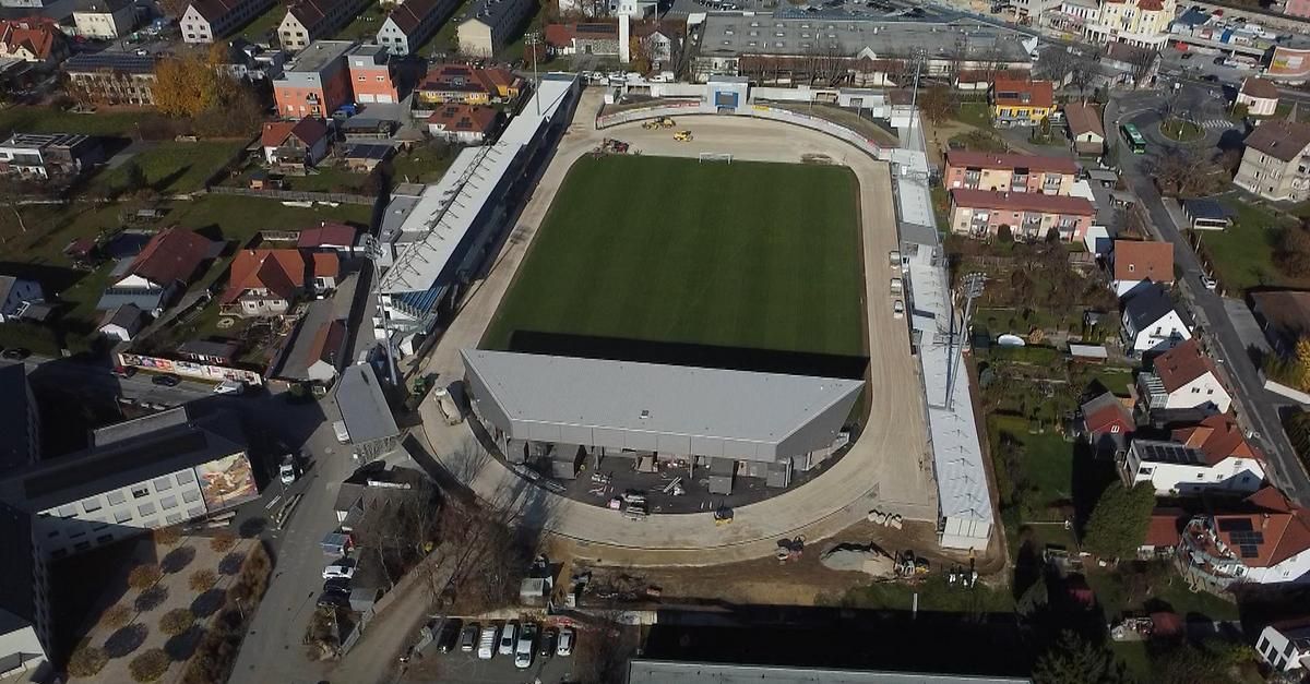 An aerial view of a sports stadium under construction with a green field, parked vehicles, and buildings in the background.