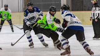 Three hockey players are on an ice rink. The player in green skates forward while two others try to block him. All wear helmets, gloves, and hockey skates.