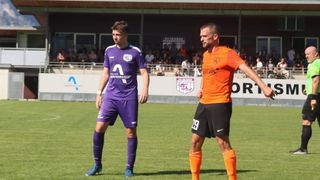 Two soccer players, one in purple and the other in orange, stand on a field. Behind them, a crowd watches from bleachers. The player in purple wears number 2.